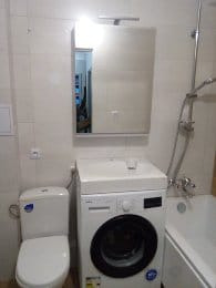 Mała łazienka z toaletą, pralką i wanną, wykończona jasnymi płytkami ceramicznymi, z szafką z lustrem nad pralką.