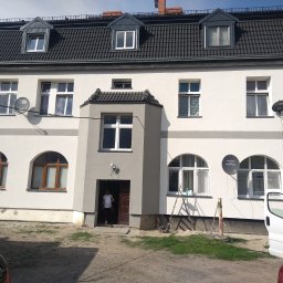 FHU Tomasz Kobyłka - Dwupiętrowy dom z szarą elewacją, ciemną dachówką i oknami o różnych kształtach, z widocznymi antenami satelitarnymi, częściowo oświetlony słońcem, z osobą wchodzącą do budynku i fragmentami...