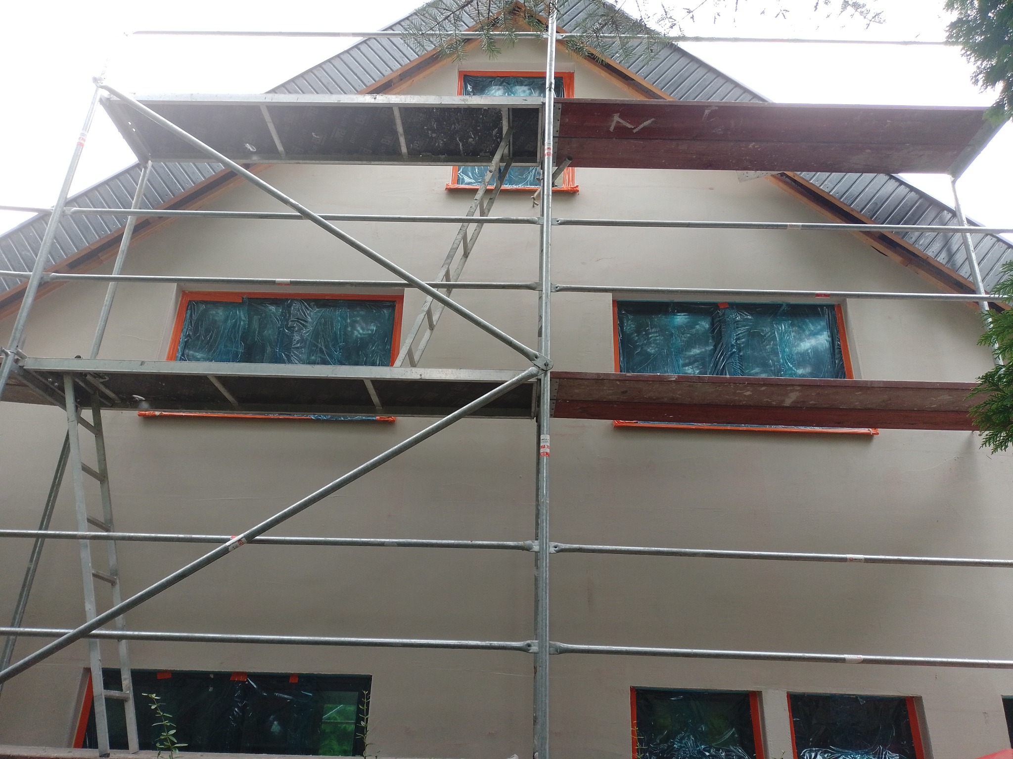 Elewacja domu w trakcie tynkowania, okna zabezpieczone folią, rusztowanie rozstawione, widoczne ślady świeżego tynku.