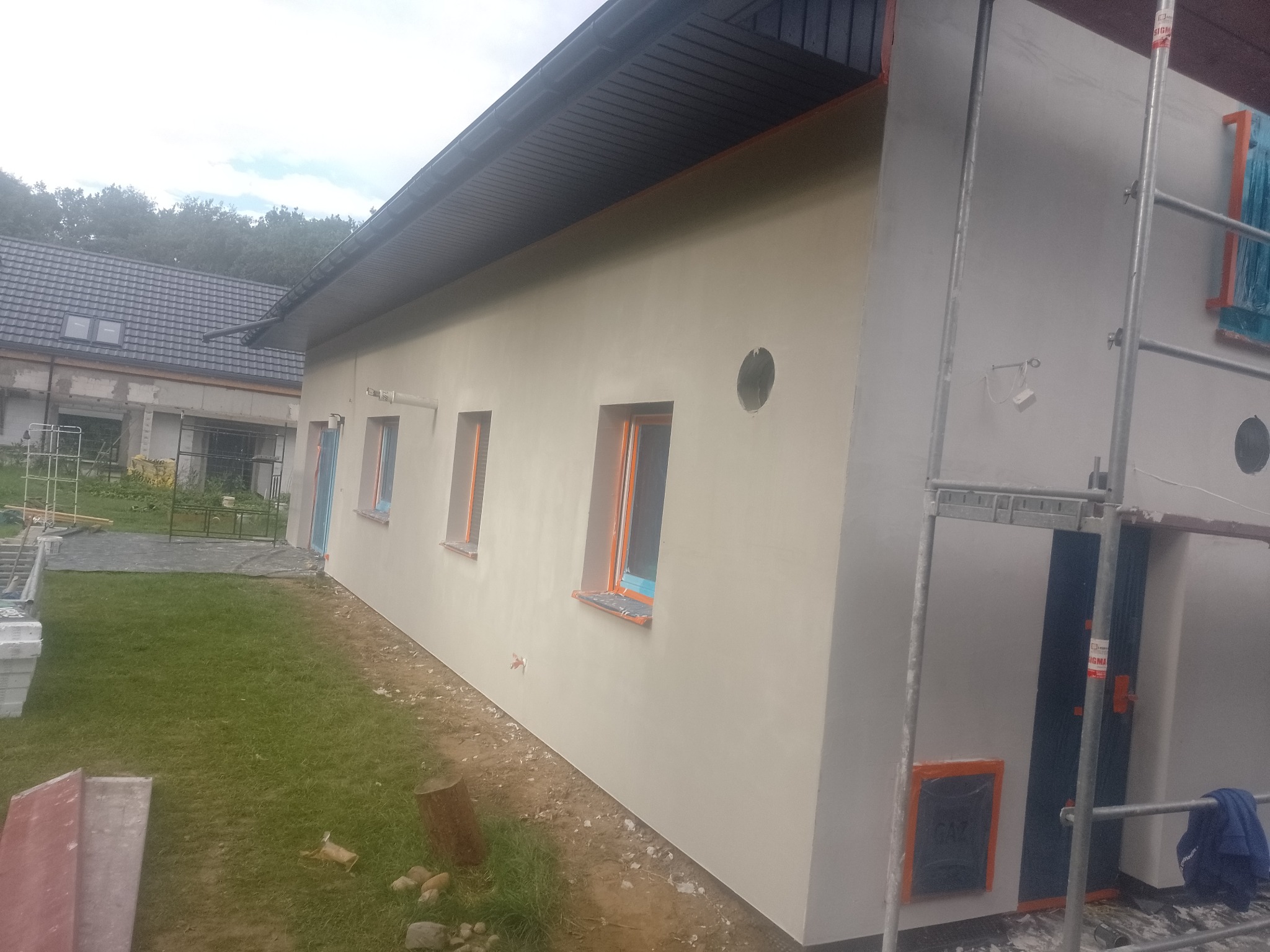 Świeżo otynkowana elewacja domu jednorodzinnego z widocznymi otworami na wentylację i oknami zabezpieczonymi folią, rusztowanie przy ścianie szczytowej, trawnik z przodu budynku.