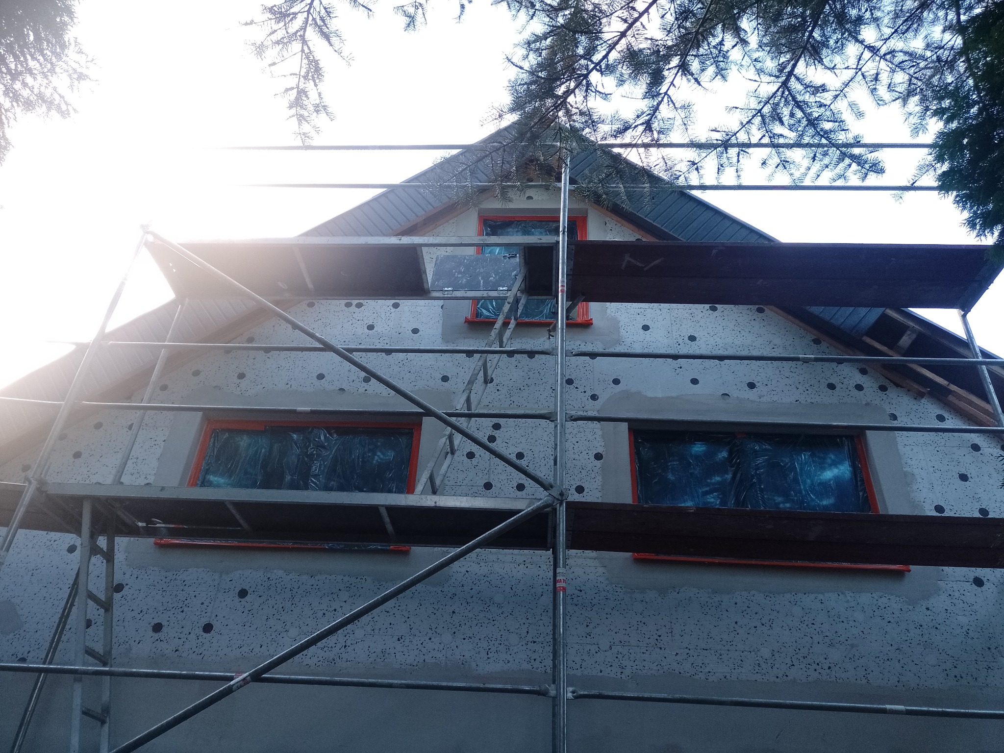 Dom w trakcie ocieplania, widoczne rusztowanie, styropian z czarnymi kołkami, okna zabezpieczone folią, fragment dachu i korony drzewa.