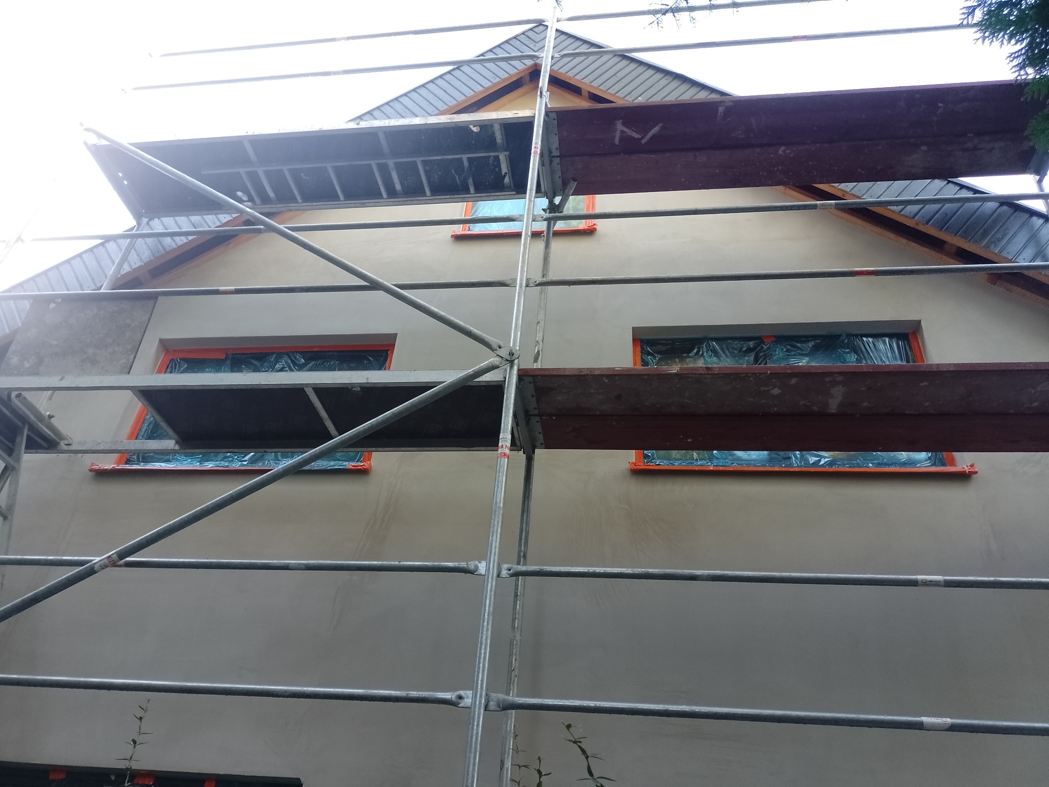 Dom jednorodzinny w trakcie tynkowania elewacji, okna zabezpieczone folią, widoczne rusztowanie.