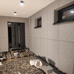 Usługi Remontowo - Budowlane Arkadiusz Kołodziej - Elewacja domu z szarymi panelami, widoczne okna i drzwi wejściowe. Na ziemi gruz i narzędzia, sugerujące prace wykończeniowe. Ujęcie z bliska, podkreślające detale.