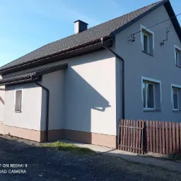 Dwukondygnacyjny dom z szarą elewacją i brązowymi rynnami, widoczny z perspektywy ogrodu, z fragmentem drewnianego płotu w pierwszym planie.