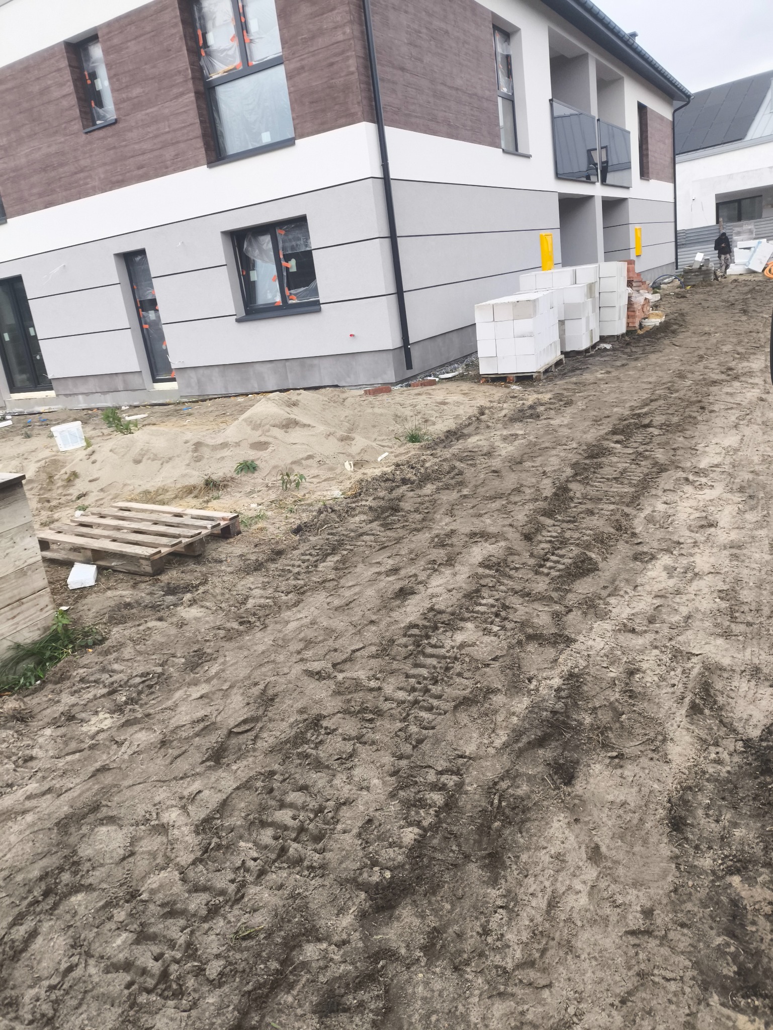 Nowoczesny budynek wielorodzinny w trakcie wykończenia elewacji, z widocznymi materiałami budowlanymi i przygotowanym terenem.