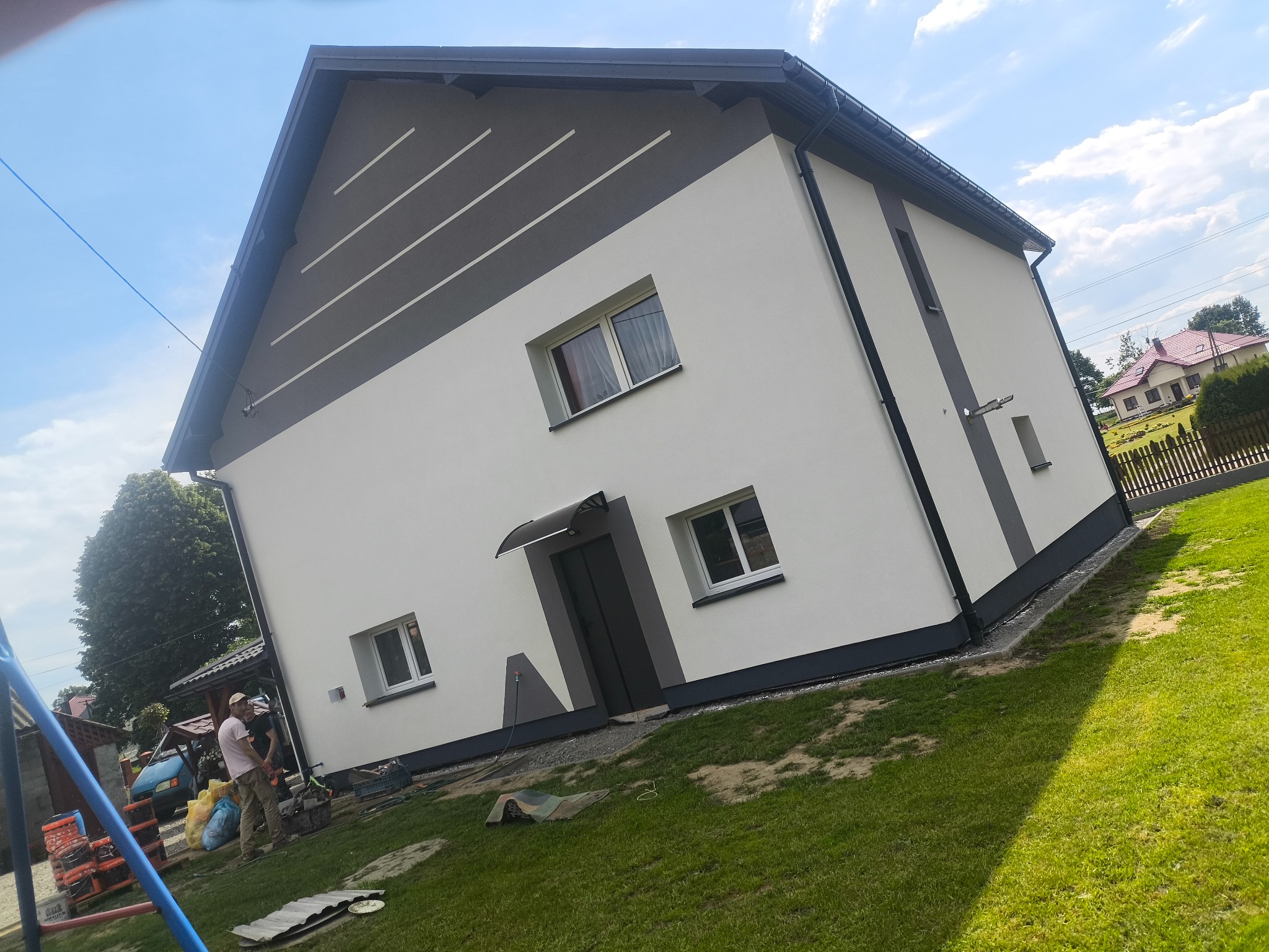 Nowoczesny dom jednorodzinny po renowacji elewacji, z białymi ścianami i szarymi akcentami. Widoczne okna, drzwi wejściowe i fragment ogrodu. Ukończony projekt.