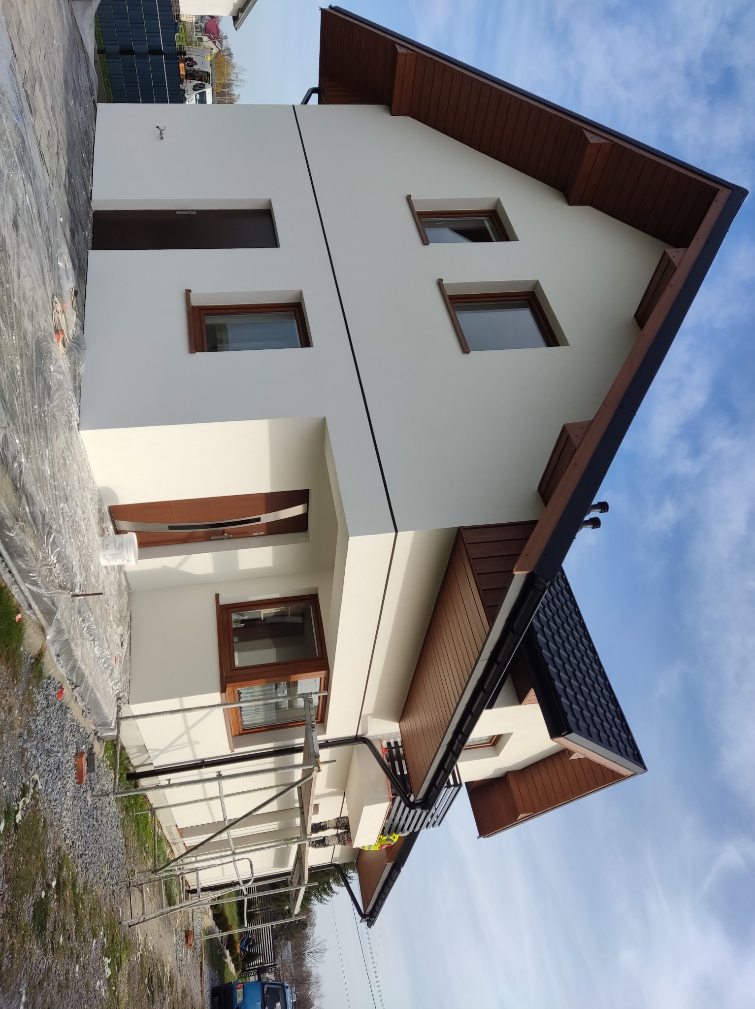 Nowoczesny dom z białą elewacją i drewnianymi elementami wykończeniowymi, widoczny z poziomu rusztowania, z niebem w tle. Część terenu zabezpieczona folią.