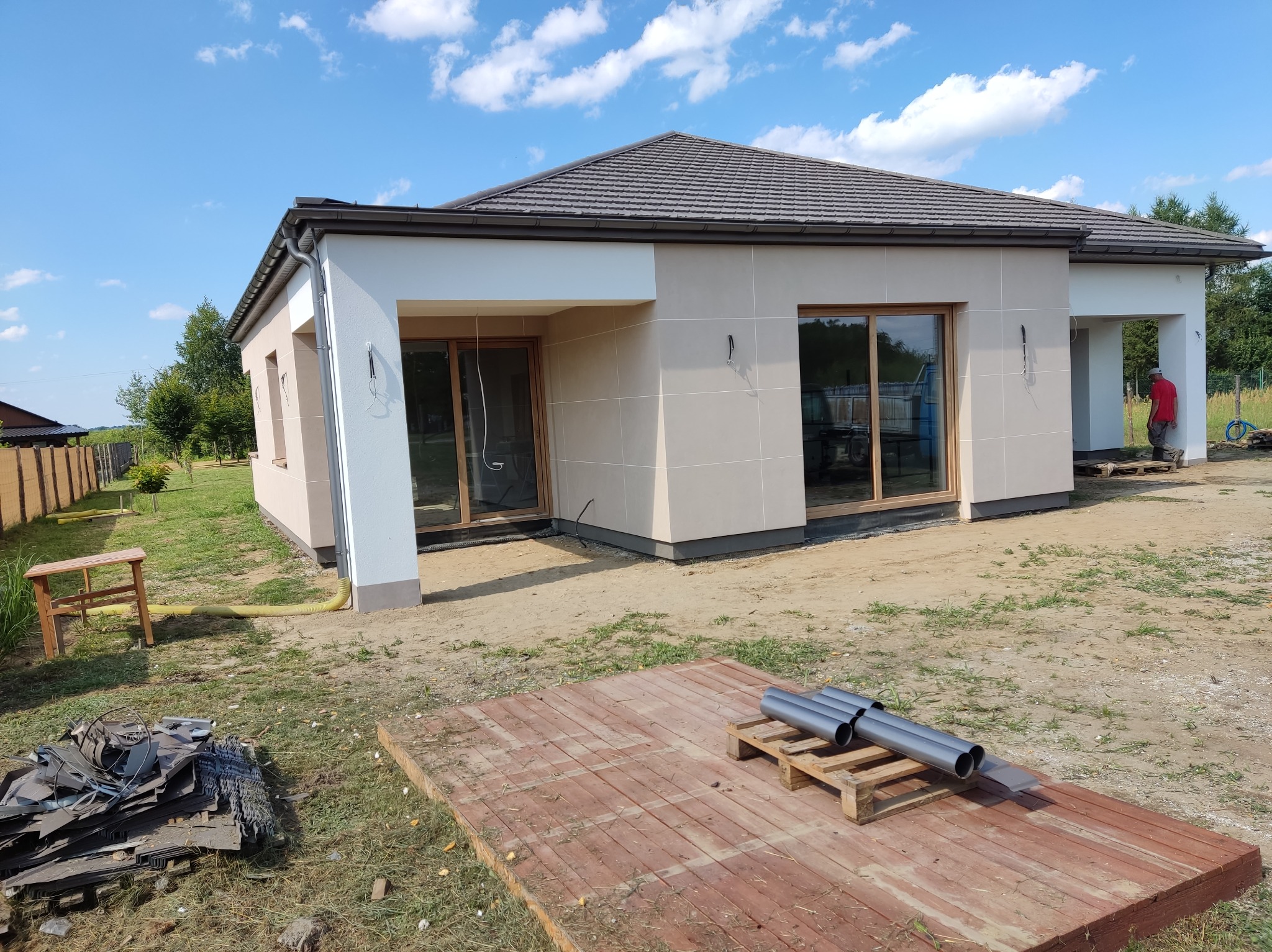 Nowoczesny dom jednorodzinny z elewacją w odcieniach beżu i bieli, z brązowym dachem. Widoczne elementy przygotowania do prac wykończeniowych na zewnątrz budynku.