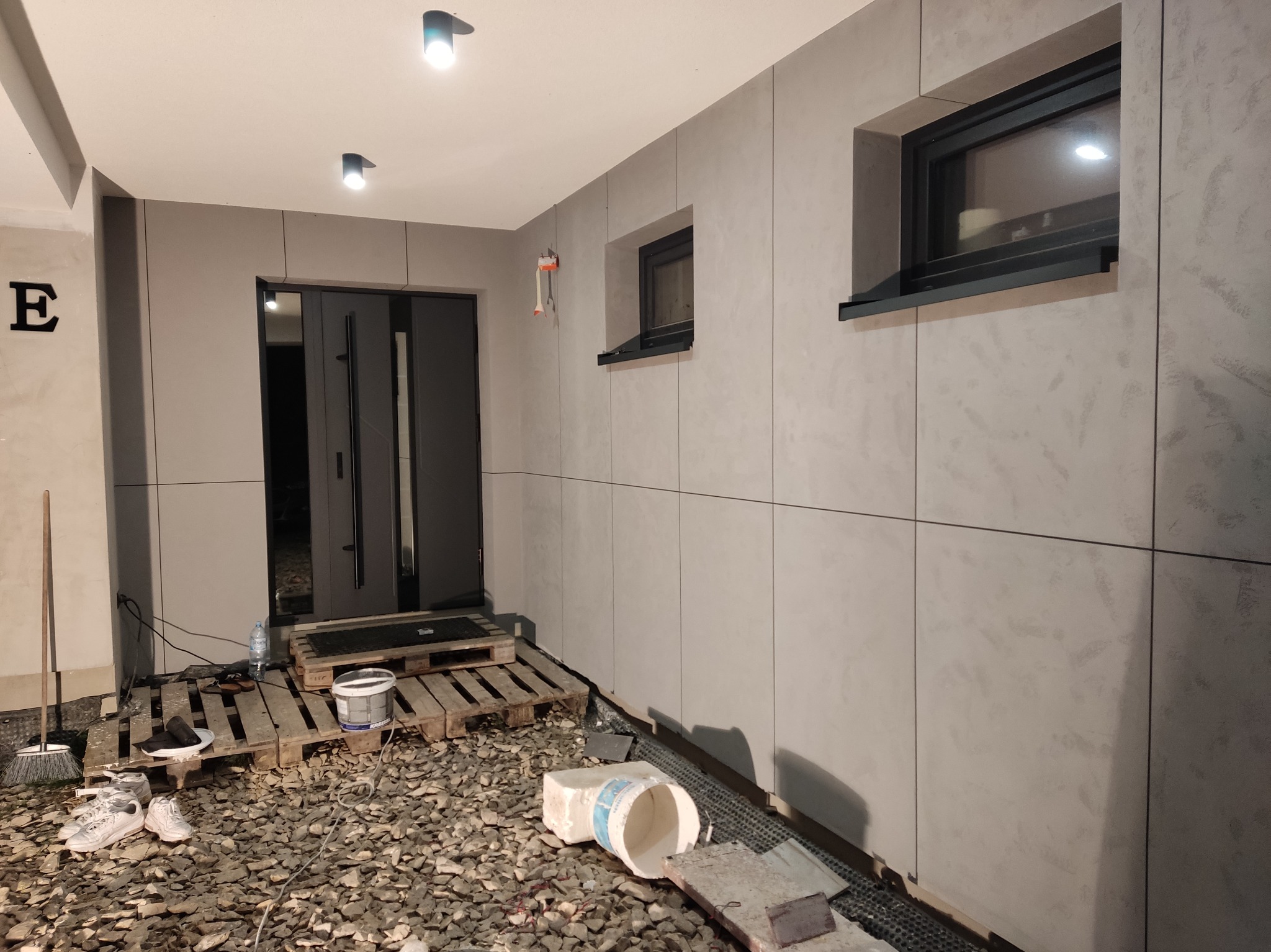 Elewacja domu z szarymi panelami, widoczne okna i drzwi wejściowe. Na ziemi gruz i narzędzia, sugerujące prace wykończeniowe. Ujęcie z bliska, podkreślające detale.