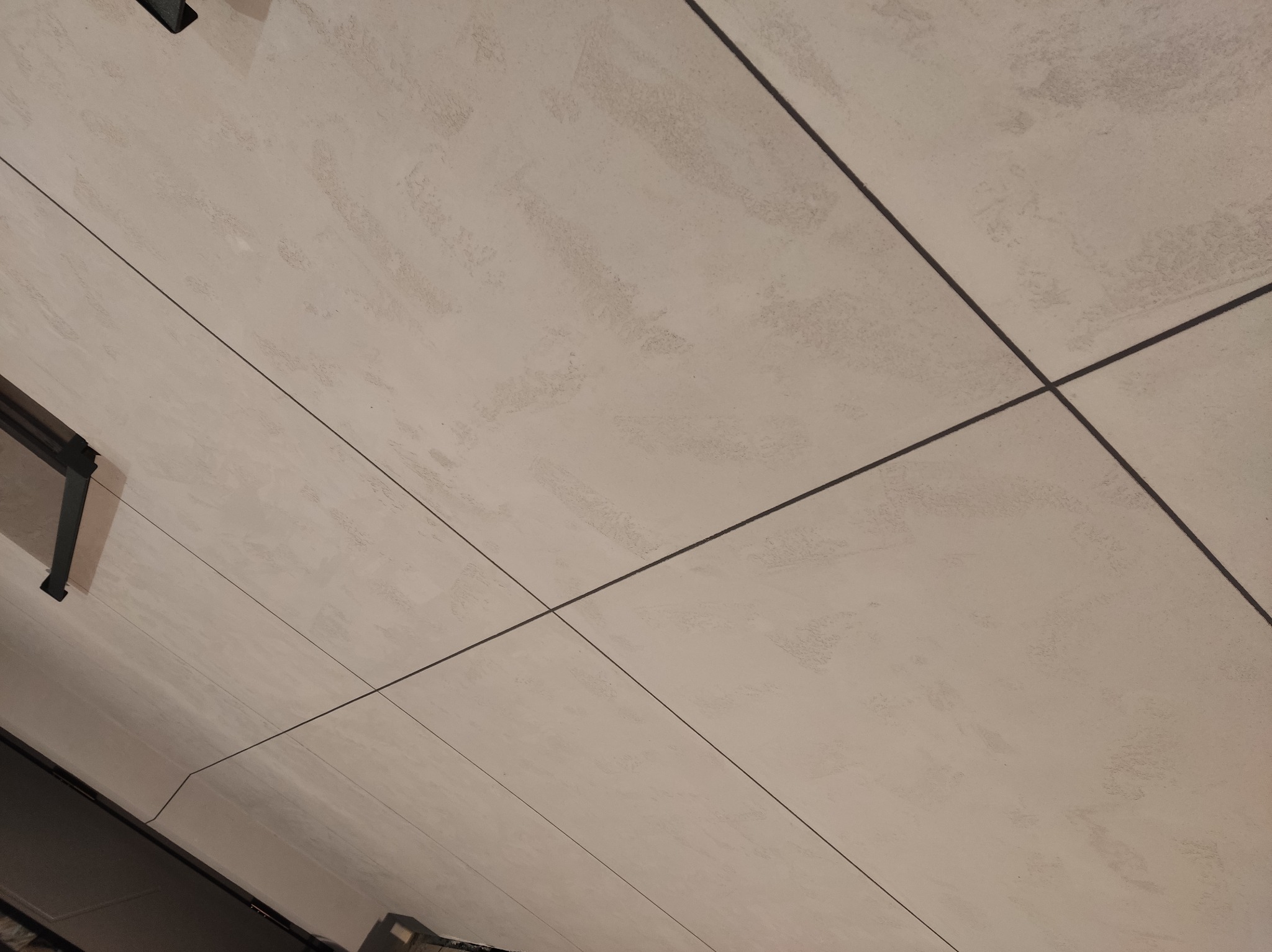 Jasna, wykończona powierzchnia sufitu z widocznymi liniami podziału płyt, sfotografowana pod kątem. Delikatna faktura i neutralna kolorystyka.