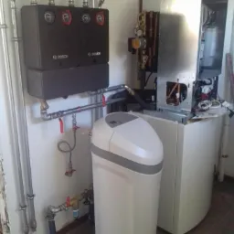 Zestaw urządzeń grzewczych Bosch z instalacją hydrauliczną, w tym zmiękczacz wody, zamontowany w kotłowni. Widoczne rury, zawory i elementy sterujące.