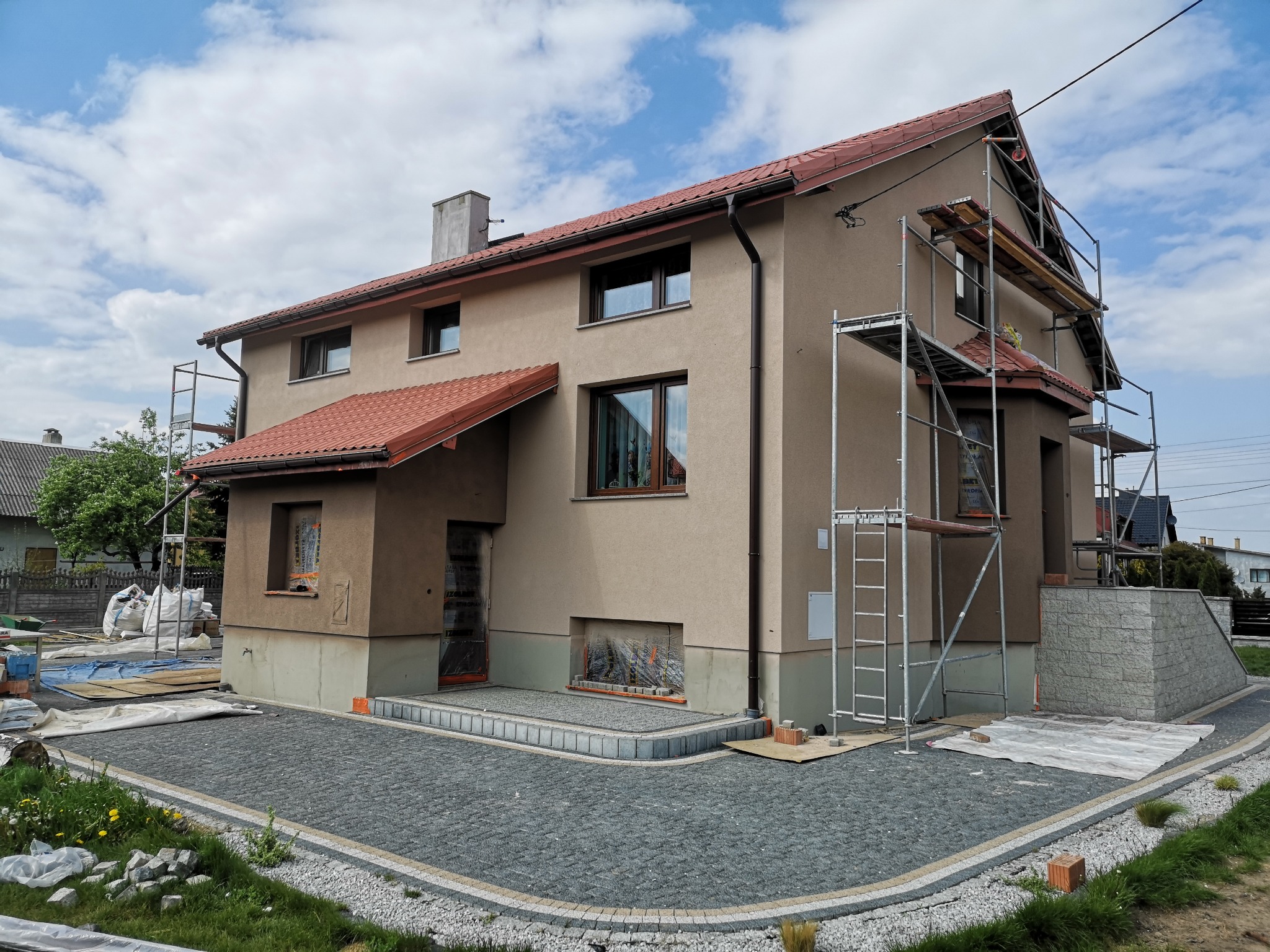 Dwupiętrowy dom w trakcie ocieplania elewacji, widoczne rusztowania i przygotowane materiały budowlane, kostka brukowa ułożona wokół domu.