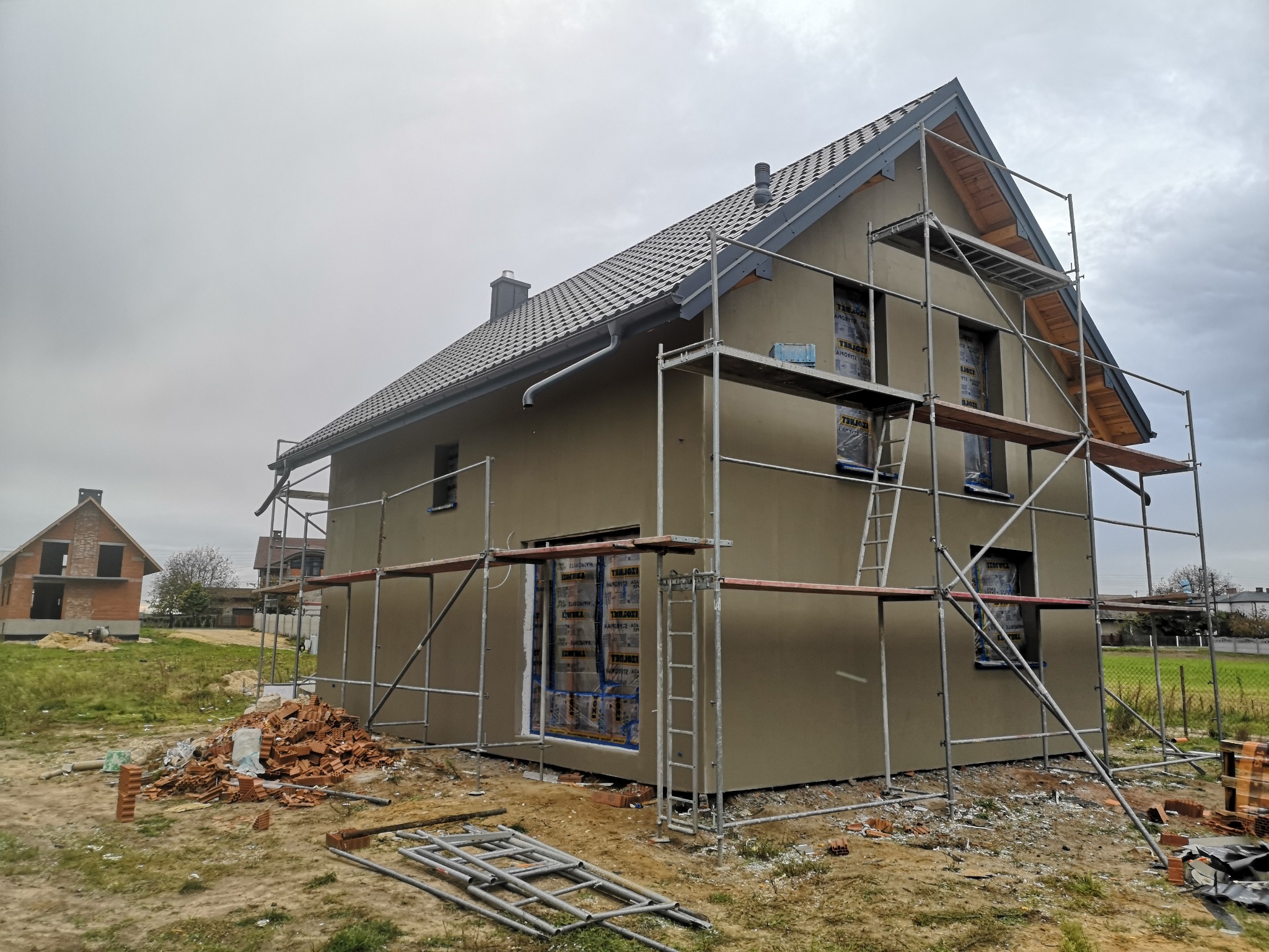 Dom jednorodzinny w trakcie prac elewacyjnych, z nałożonym tynkiem i ustawionymi rusztowaniami, okna zabezpieczone folią, na pierwszym planie sterta cegieł i fragmenty rusztowań.