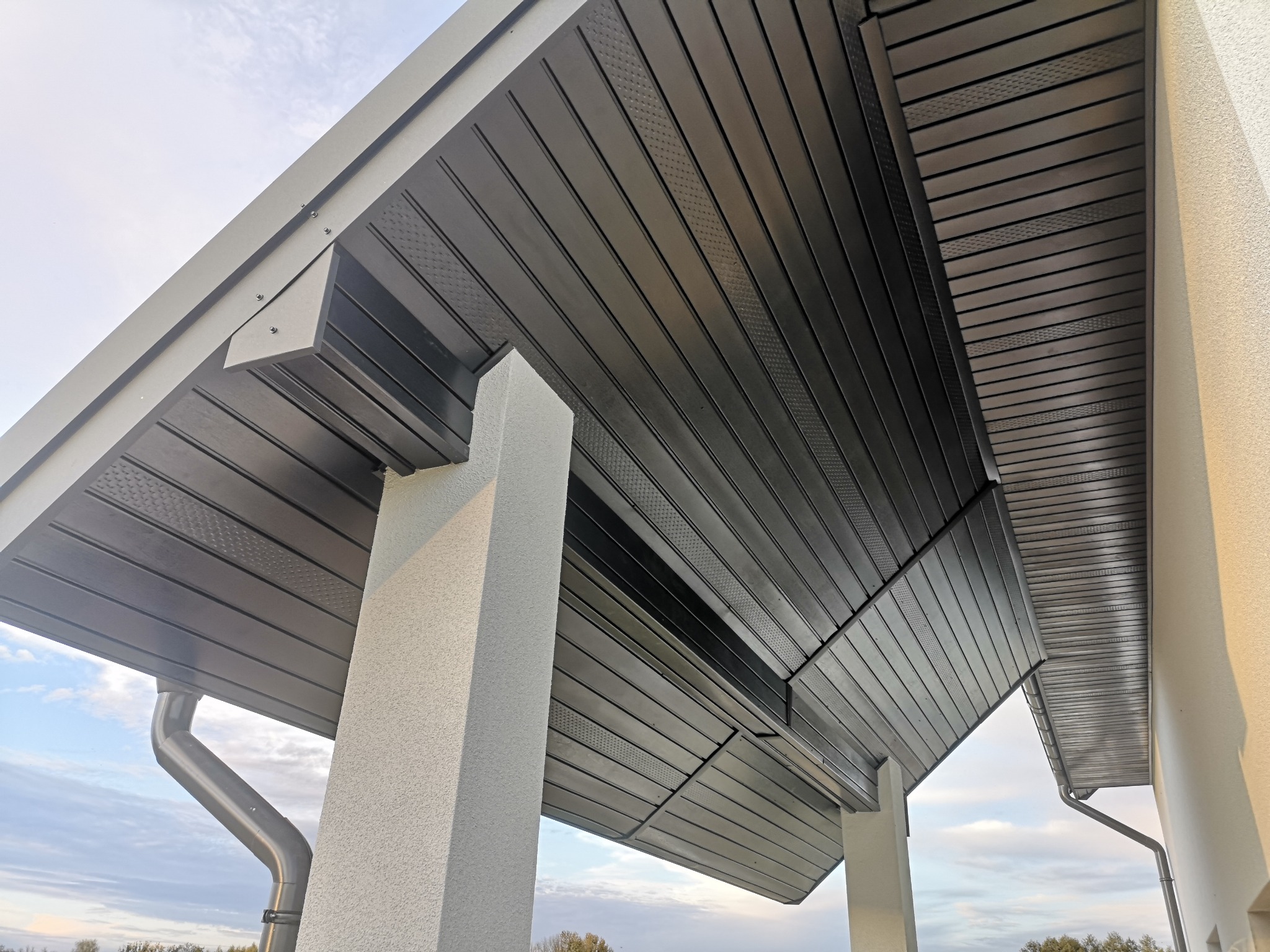 Nowoczesna podbitka dachowa w kolorze antracytowym, wykonana z paneli z delikatnym wzorem, zamontowana przy budynku z jasną elewacją i widoczną rynną spustową.