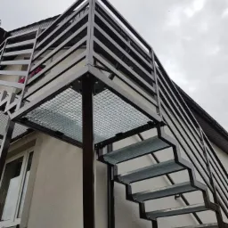 Zewnętrzne schody stalowe z platformą, ocynkowane ogniowo, z czarną balustradą, przymocowane do budynku z oknem.