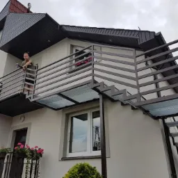 Zewnętrzne schody stalowe i balkon z metalową balustradą przy dwukondygnacyjnym domu z kwiatami w oknach, widok z dołu.