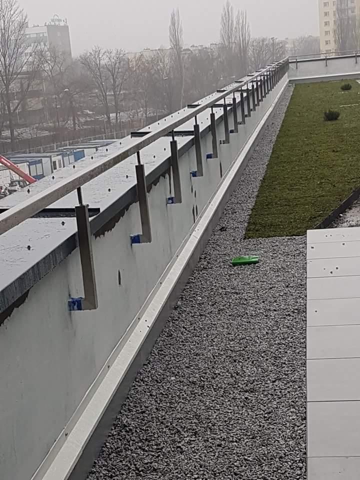 Montaż stalowej balustrady na dachu budynku z zielonym dachem w tle, widoczne elementy mocujące i żwir.