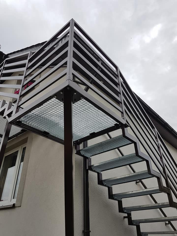 Zewnętrzne schody stalowe z platformą, ocynkowane ogniowo, z czarną balustradą, przymocowane do budynku z oknem.