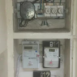 Wnęka z trzema licznikami elektrycznymi o różnych modelach i numerach seryjnych, zamontowanymi na surowej, pomalowanej ścianie, powyżej widoczna skrzynka z bezpiecznikami topikowymi i przewodami...