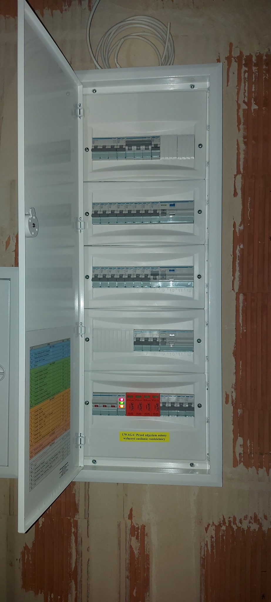 Otwarta skrzynka rozdzielcza z bezpiecznikami na ścianie w trakcie montażu w Gliwicach, widoczne przewody i etykieta ostrzegawcza.