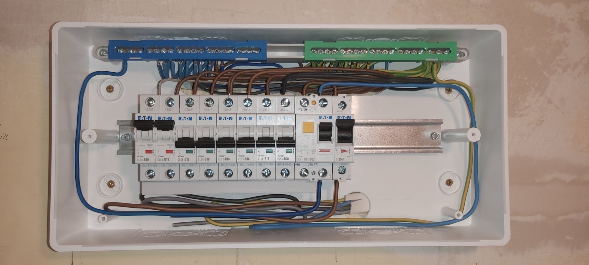 Rozdzielnica elektryczna Eaton z zainstalowanymi bezpiecznikami B10, B16 i B20, widoczne niebieskie i zielone listwy zaciskowe oraz przewody w różnych kolorach, zamontowana w białej obudowie na tle...