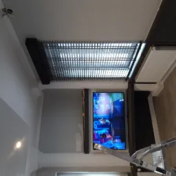 Widok z dołu na zamontowany telewizor na ścianie, obok grzejnik, na suficie kratka wentylacyjna, widoczna drabina teleskopowa.