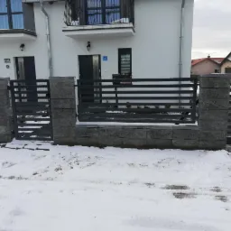 Nowoczesne ogrodzenie metalowe z elementami z szarego kamienia łupanego na tle ośnieżonego podjazdu przed białym domem.