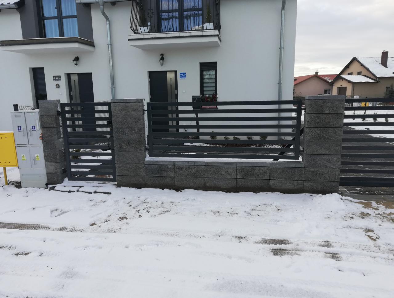Nowoczesne ogrodzenie metalowe z elementami z szarego kamienia łupanego na tle ośnieżonego podjazdu przed białym domem.