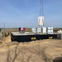 Budowa domu: dźwig podnosi bloczki Solbet nad fundamentami, widoczni robotnicy w trakcie prac murarskich, w tle krajobraz z polami i błękitne niebo.