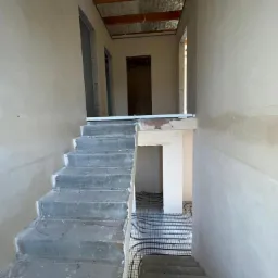 Wnętrze domu w trakcie prac wykończeniowych: surowy beton schodów, ściany po nałożeniu gładzi oraz instalacja ogrzewania podłogowego na siatce w dolnej części pomieszczenia.