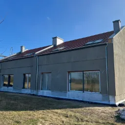 Surowy stan domu szeregowego z szarym tynkiem, czerwoną dachówką i oknami bez wykończenia, widoczne rynny i kominy, teren wokół w trakcie zagospodarowania.