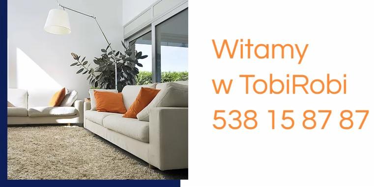 Jasny salon z beżowym dywanem, kremowymi sofami z pomarańczowymi poduszkami, lampą łukową i dużym oknem, obok pomarańczowy napis 'Witamy w TobiRobi 538 15 87 87'.