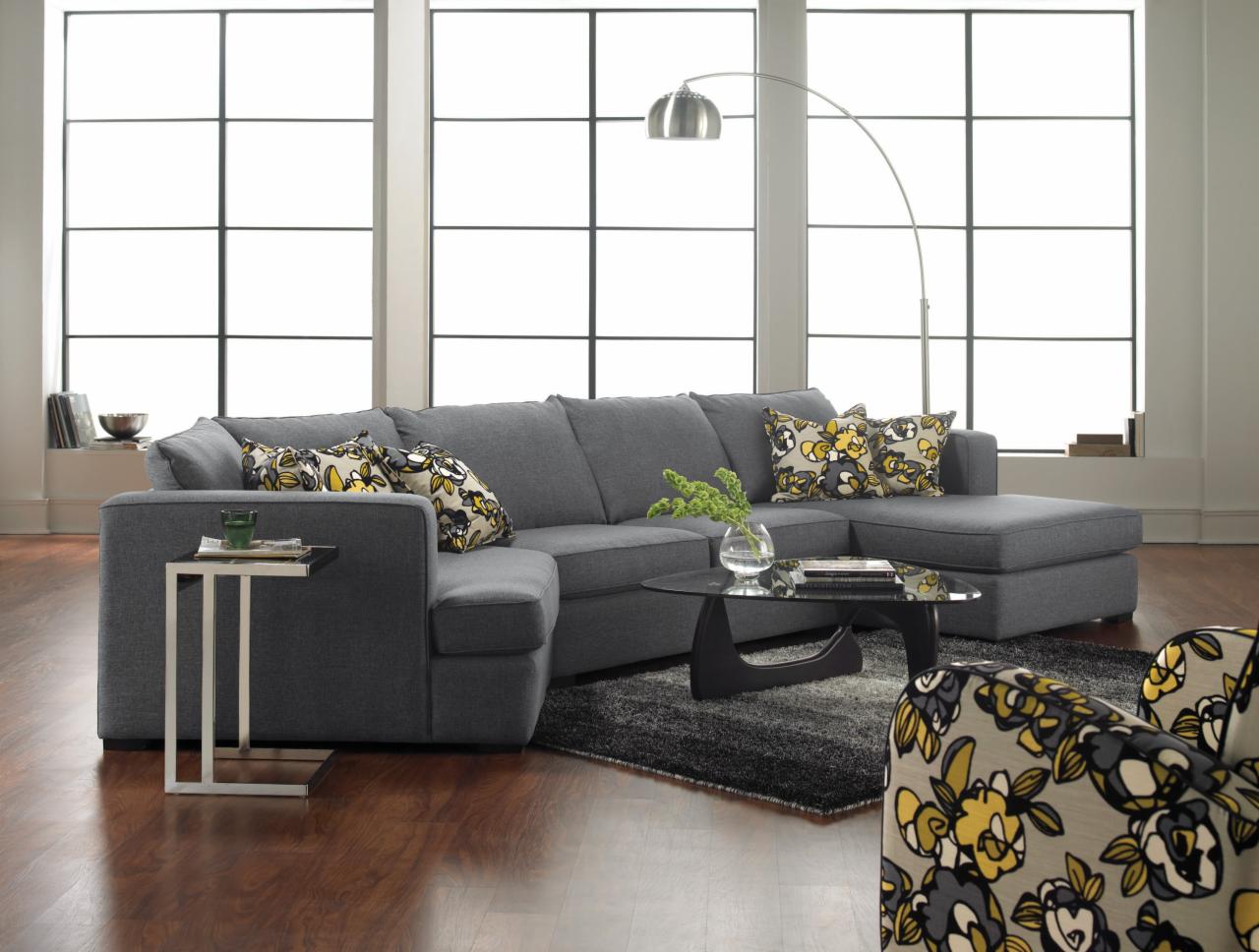 Szara, modułowa kanapa narożna z szezlongiem, wzorzyste poduszki i fotel z kwiatowym motywem na tle dużego okna i drewnianej podłogi.