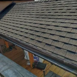 Nowy dach pokryty gontem bitumicznym, widoczny z rusztowania; brązowa rynna zamontowana wzdłuż krawędzi dachu.