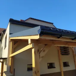 Drewniana konstrukcja z zadaszeniem z blachy trapezowej przy budynku, widoczne rynny w kolorze czarnym, błękitne niebo w tle.