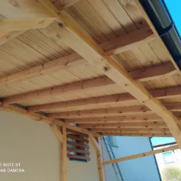 Drewniana konstrukcja z widoczną więźbą dachową, deskowaniem i rynną w kolorze czarnym, sfotografowana z dołu na tle błękitnego nieba.