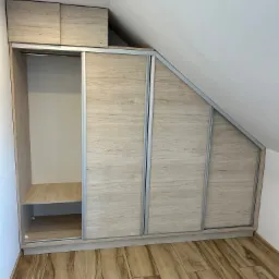 Drewnopodobna szafa wnękowa z przesuwnymi drzwiami, dopasowana do skosu dachu, z widoczną półką i drążkiem na ubrania.