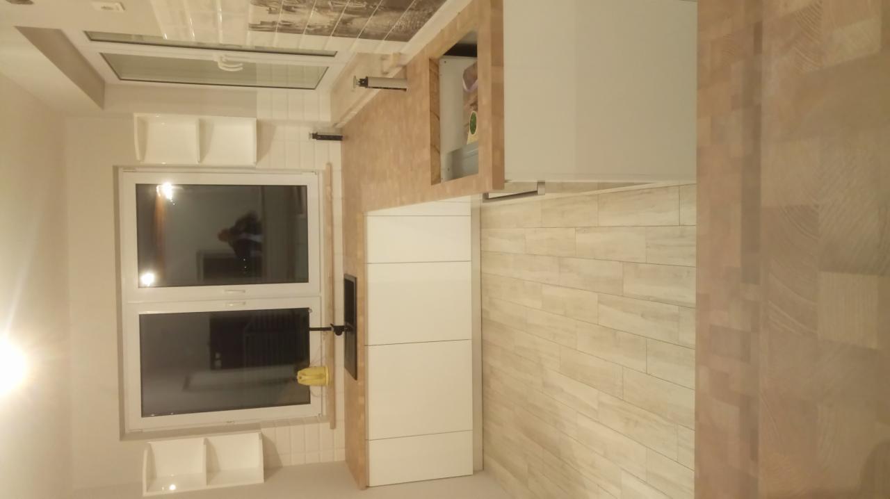 Nowoczesna kuchnia z białymi frontami szafek i drewnianym blatem, widok na okno z półkami i jasne płytki podłogowe imitujące drewno.