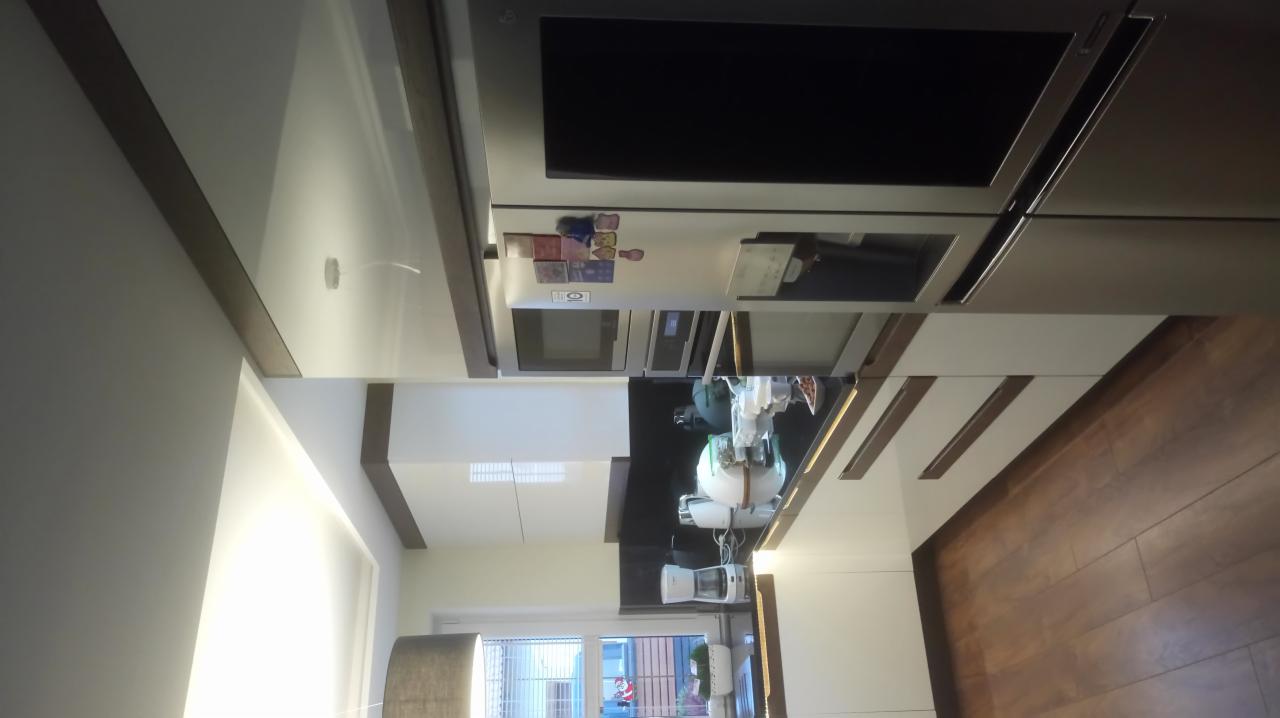 Nowoczesna kuchnia z białymi, lakierowanymi frontami, drewnianym blatem i zabudowanym sprzętem AGD. Widoczny ekspres do kawy i inne akcesoria kuchenne. Ujęcie z perspektywy sufitu.