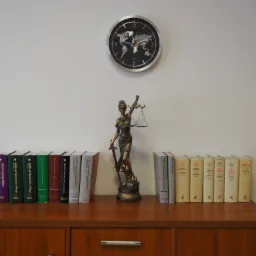 Statuetka Temidy z wagą i mieczem, otoczona książkami prawniczymi na drewnianej komodzie, nad nią zegar z mapą świata.