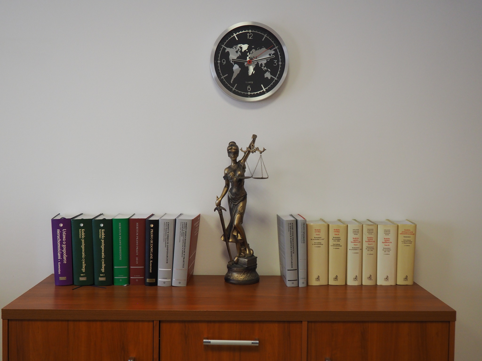 Statuetka Temidy z wagą i mieczem, otoczona książkami prawniczymi na drewnianej komodzie, nad nią zegar z mapą świata.