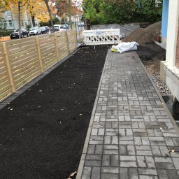 Usługi brukarskie i budowlano remontowe - Ułożona szara kostka brukowa tworząca ścieżkę obok świeżo nawiezionej ziemi pod trawnik, oddzielonej jasnym drewnianym płotem, w tle budynek w trakcie wykończenia.