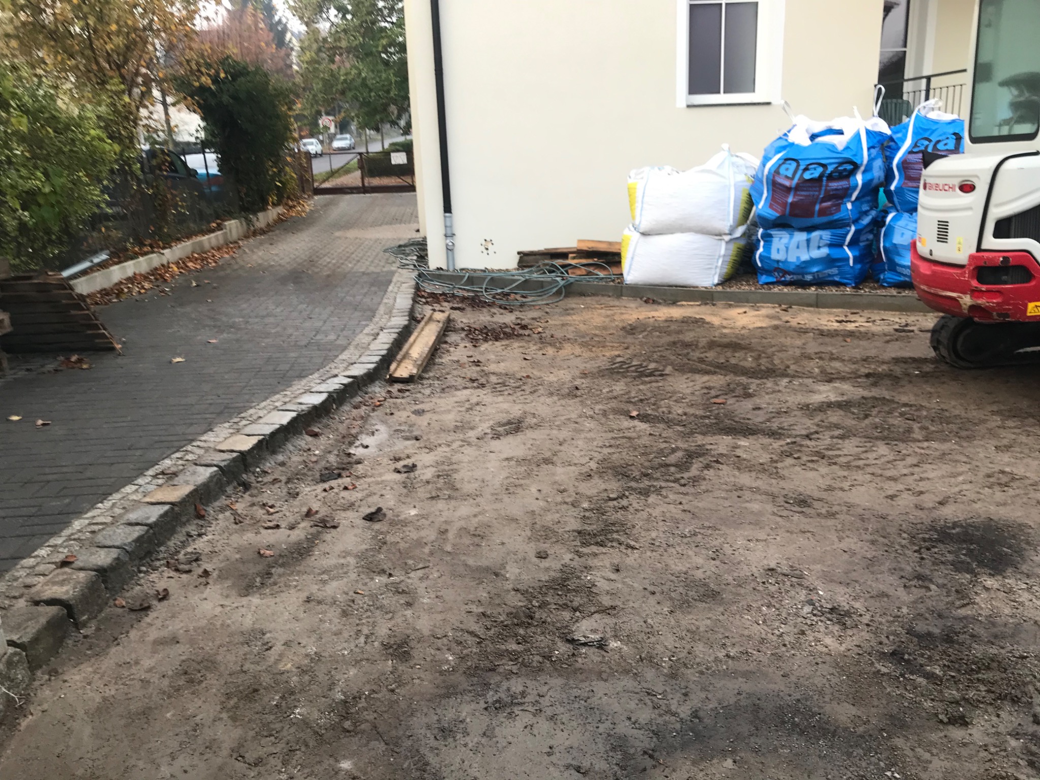 Przygotowanie terenu pod kostkę brukową: widoczne worki z materiałem, mini koparka oraz krawężnik oddzielający chodnik od przyszłej nawierzchni.