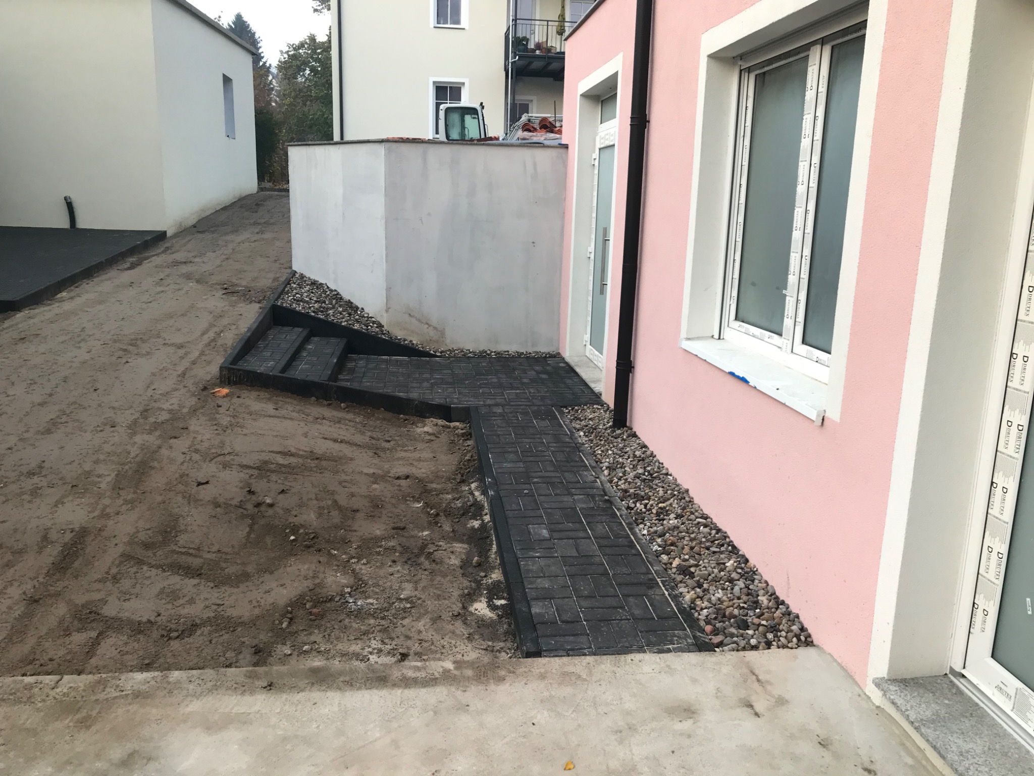 Ścieżka z ciemnej kostki brukowej prowadząca do różowego budynku, z widocznymi schodami i obsypanym kamieniami obszarem wzdłuż ściany, teren wokół w trakcie prac ziemnych.