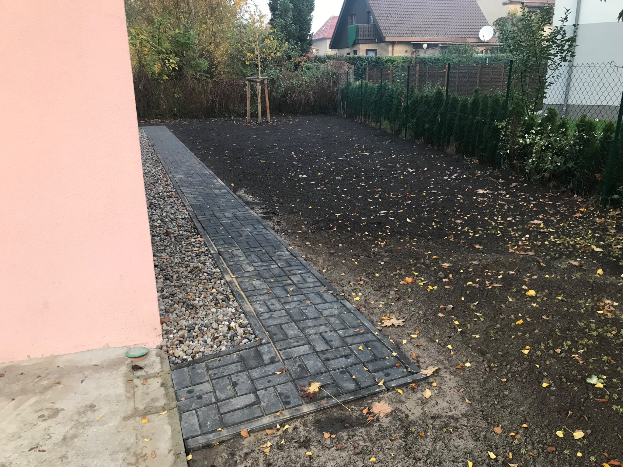 Świeżo ułożona, ciemnoszara kostka brukowa prowadząca wzdłuż różowej ściany budynku, obok pas jasnych kamyków, przygotowany teren pod trawnik w tle.