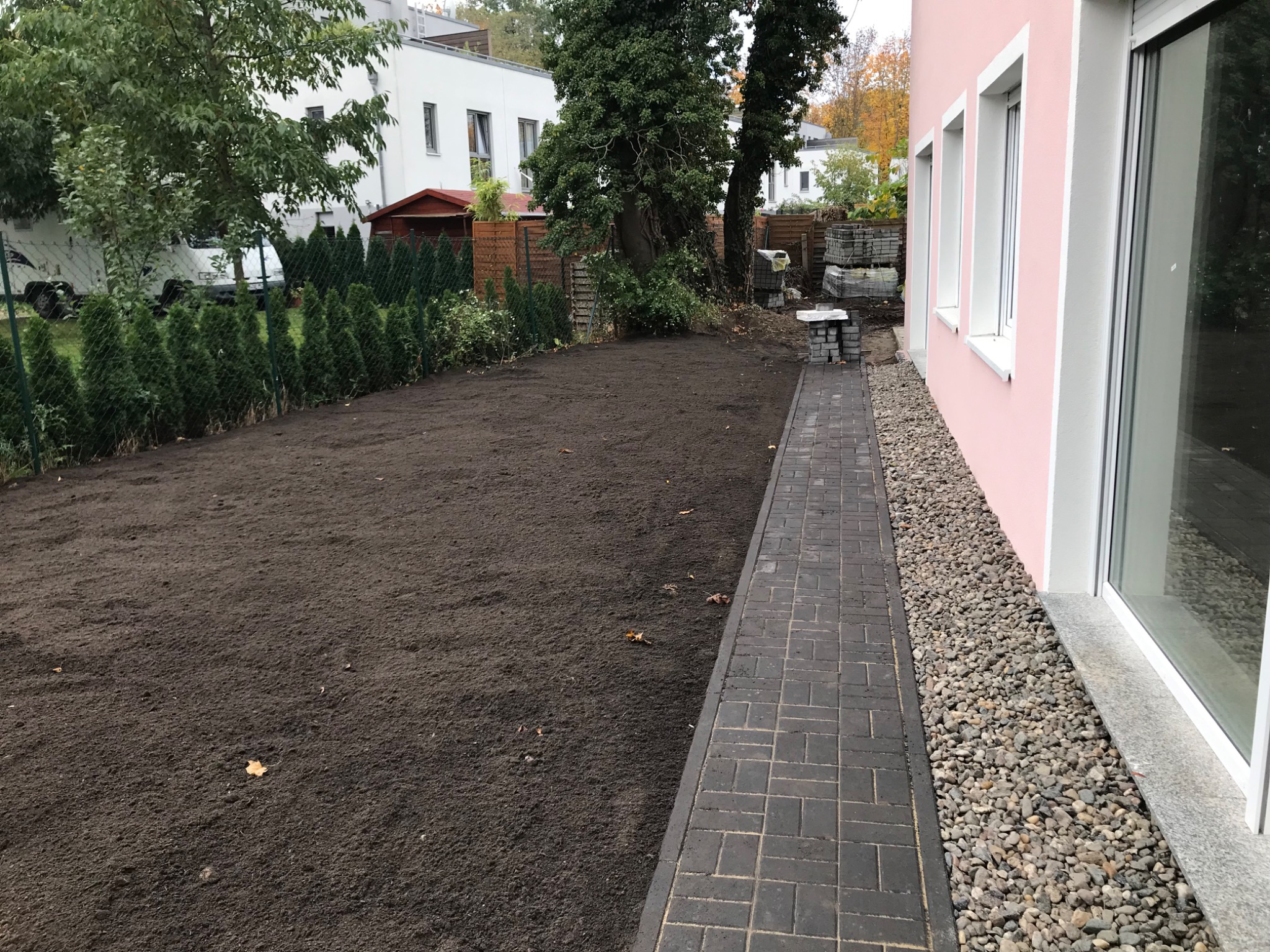 Ułożona ścieżka z ciemnoszarej kostki brukowej wzdłuż różowego budynku, świeżo wyrównana ziemia pod trawnik, ułożone kostki do dalszych prac, widok na dom i drzewa w tle.