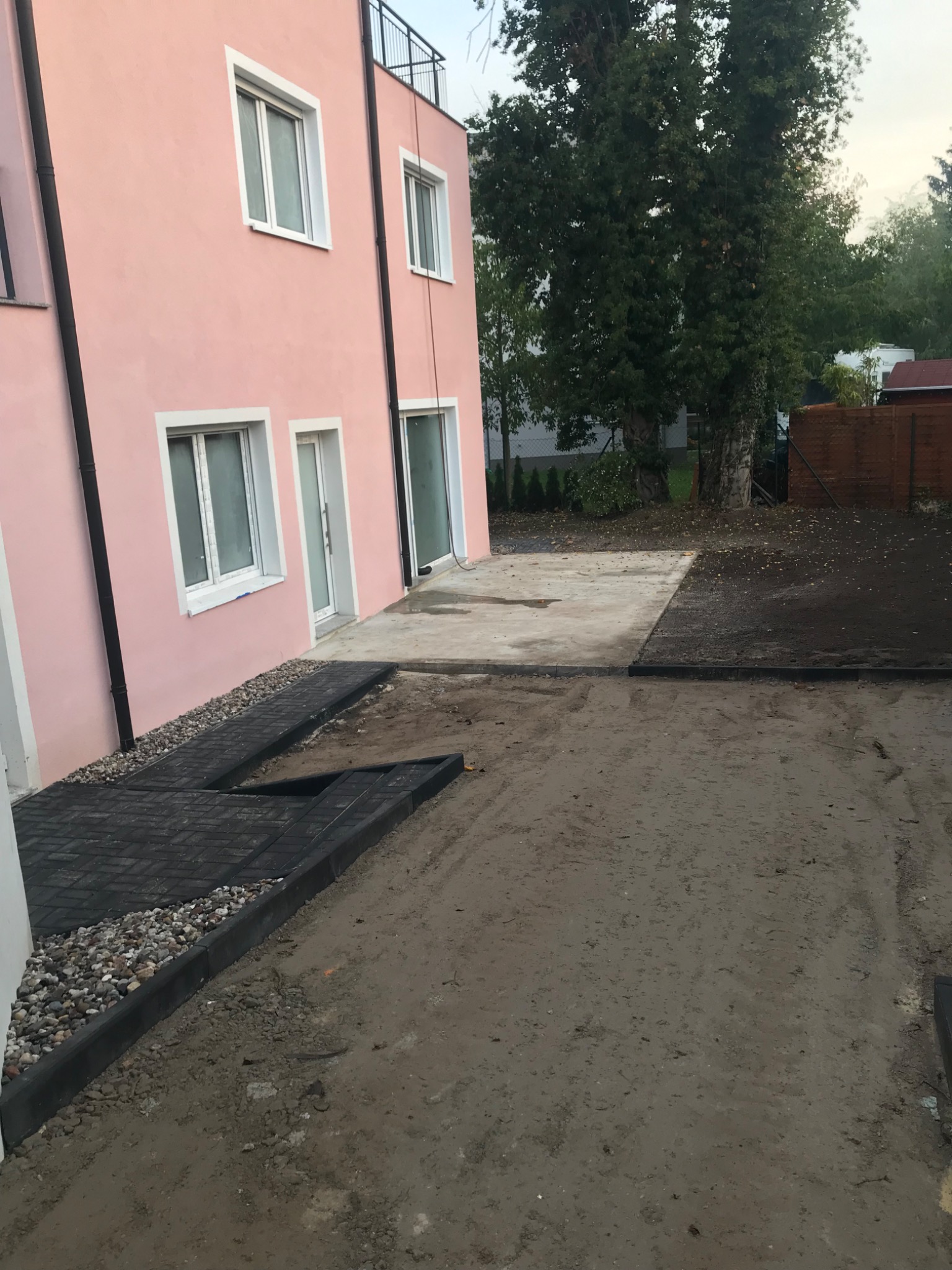 Częściowo ułożona ścieżka z ciemnej kostki brukowej prowadząca do wejścia budynku o różowej elewacji, widoczne podłoże przygotowane pod dalsze prace.