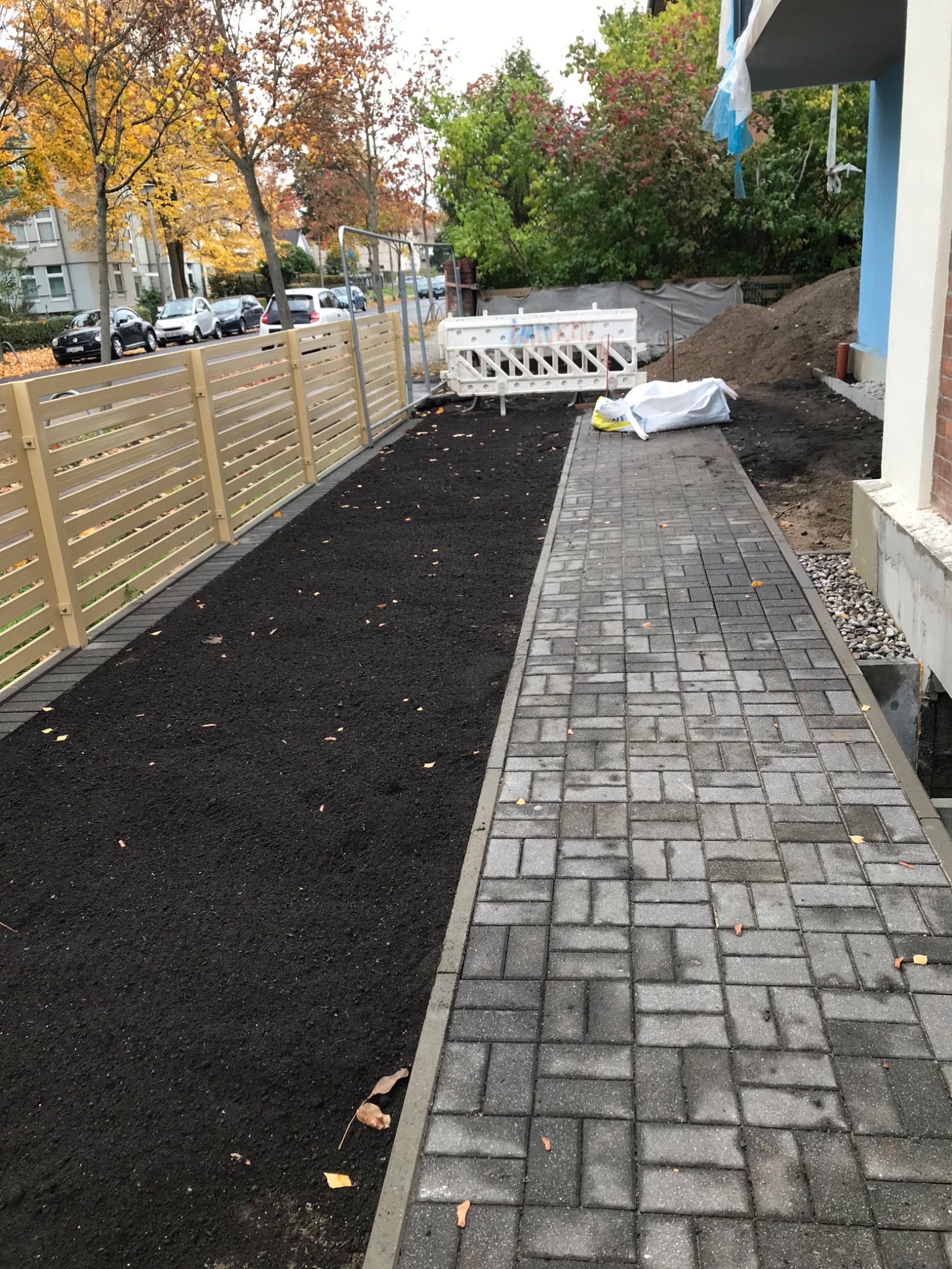 Ułożona szara kostka brukowa tworząca ścieżkę obok świeżo nawiezionej ziemi pod trawnik, oddzielonej jasnym drewnianym płotem, w tle budynek w trakcie wykończenia.