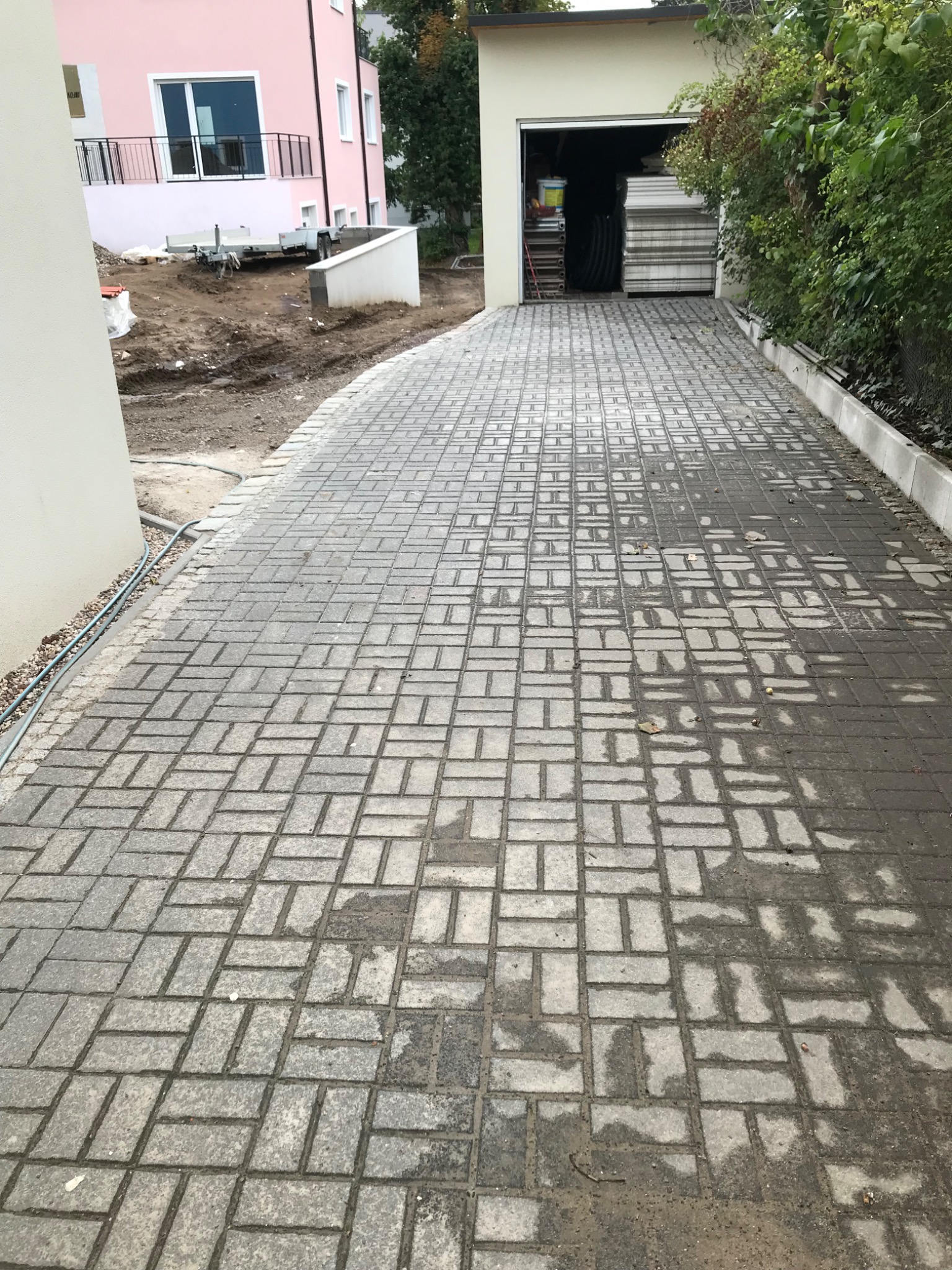 Ułożona szara kostka brukowa na podjeździe do garażu, widoczne ślady wilgoci i niedokończone prace ziemne obok różowego domu.