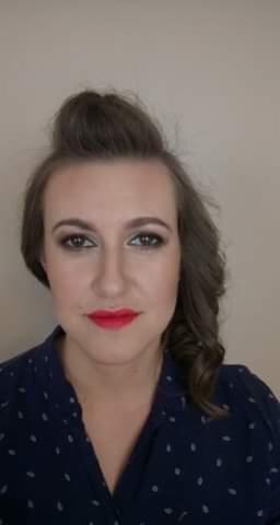 Portret kobiety z upiętymi włosami, wyrazistym makijażem oczu w odcieniach szarości i intensywnie czerwoną szminką, ubrana w granatową koszulę w białe kropki.